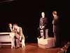 Four people are engaged in a theatrical scene on a dimly lit stage. Two are sitting at a table on the left, one is seated on a raised platform center stage, and the fourth stands to the right. Their expressions appear serious.