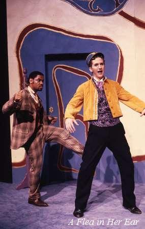 Two actors perform on stage. One wears a plaid suit and gestures dramatically, while the other wears a yellow jacket and blue hat, mid-stride with a surprised expression. A colorful, abstract door design is in the background. "A Flea in Her Ear" text present.