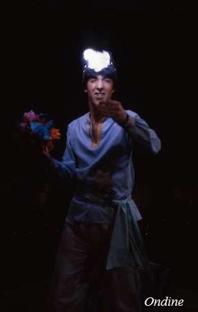 A person wearing a light blue shirt and pants, holding a colorful bouquet, and pointing forward. Their head is illuminated by a bright light, creating a dramatic effect. The background is dark, making the figure and the light stand out prominently.