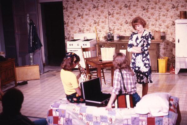 A woman in a floral dress stands in a kitchen with floral wallpaper, observing two girls sitting on a bed. One girl wears a yellow top and hairband, and the other wears a plaid shirt. They're focused on an open case containing a chess set.