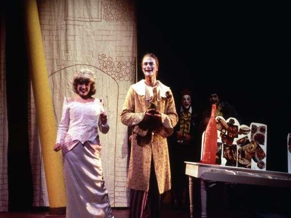 Two actors in theatrical makeup and costumes perform on stage. One wears a white dress, and the other a long coat with patterns. A table with large props, including a red bottle, is to the right. A painted backdrop and other figures are in the background.
