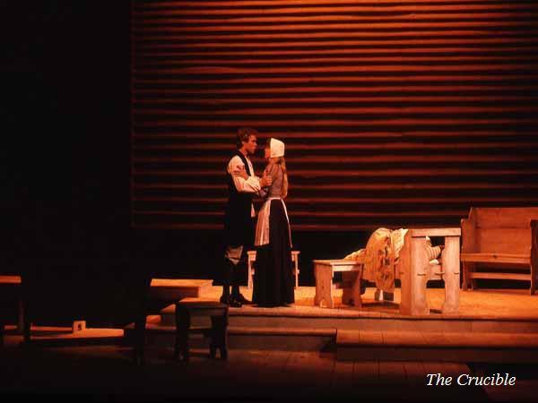 A dimly lit stage of a theater production shows two actors in period costumes. The set resembles a rustic interior with wooden furniture. The caption reads "The Crucible" in the lower right corner.