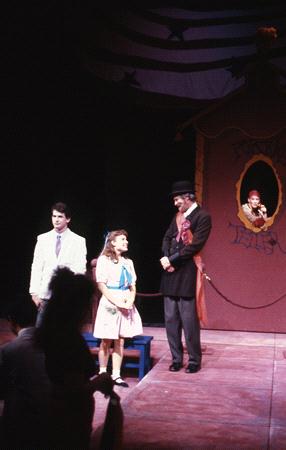 Three actors performing on stage in a play, with one wearing a white suit, a child in a colorful dress, and another in a black suit with a top hat. A fourth actor appears in a booth in the background. Stage lights highlight the scene.