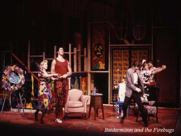A stage performance of "Biedermann and the Firebugs" featuring actors in colorful costumes interacting animatedly. The set includes a chair, floral wreath, and abstract background paintings, creating a lively theatrical scene.