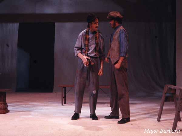 Two actors on stage perform a scene from a play. One wears a scarf and hat, holding an object in hand, while the other wears a cap and vest. The set includes simple wooden benches. The backdrop is minimalistic. Text reads "Major Barbara.