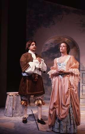 A man and woman in period costumes perform on stage. The man wears a brown outfit with puffy sleeves and stockings. The woman wears a flowing pink dress. They stand in front of a backdrop with an arch design.