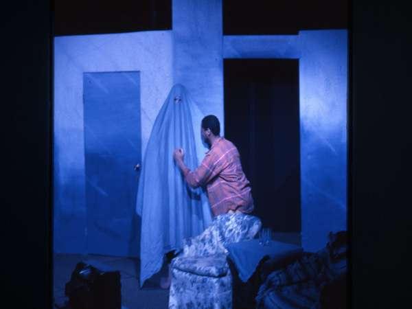 A person kneels and gestures toward a figure covered by a blue sheet, resembling a ghost. The scene is set in a dimly lit room with blue-toned walls and furniture.