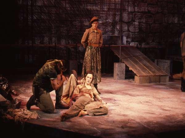 Three people are on a stage. One person lies on the ground, another kneels beside them, and a third stands in the background. The setting is dark, with an earthy, rustic backdrop. A ramp leads off the stage in the background.