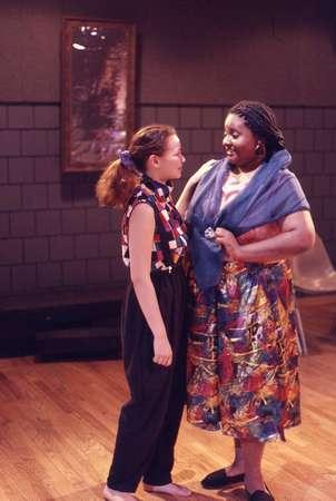 Two people stand in a room with wooden floors and dark brick walls. One wears a colorful skirt and scarf, and the other wears a black and red patterned outfit. They are holding hands and smiling at each other. A framed picture hangs on the wall behind them.