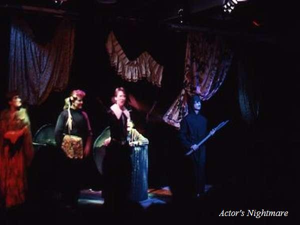 A dimly lit stage with four performers in costume. They stand in front of dramatic, draped fabric. The scene has a theatrical, mysterious atmosphere. "Actor's Nightmare" is written in the bottom right corner.
