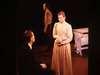 A woman in a long skirt and light-colored blouse stands facing a seated person in dark clothing. Another person stands in the background near a bed. The scene appears to be set on a stage.