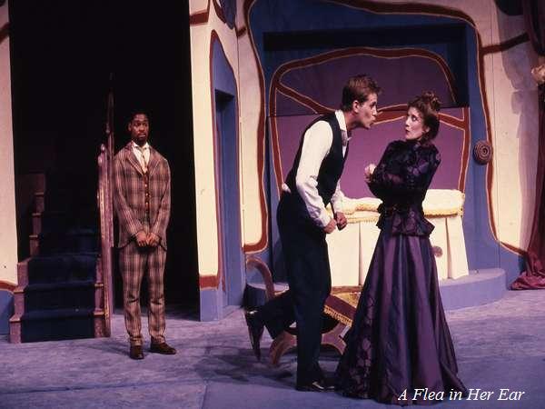 Three actors performing on stage. A man in a plaid suit stands left, while a man in a vest angrily talks to a woman in a purple dress. The background has abstract purple and beige patterns. The text "A Flea in Her Ear" is in the bottom right corner.