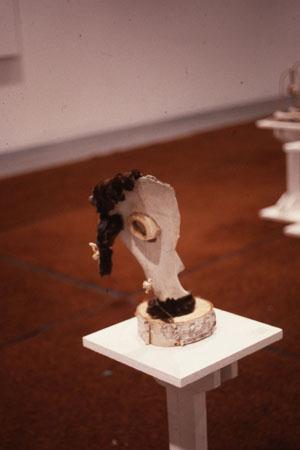Abstract sculpture resembling a side profile of a face, crafted from organic materials, with a prominent wood texture. The piece is displayed on a small white pedestal, set against a brown carpeted floor.