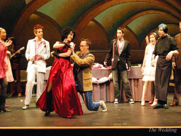 A theater scene where a person in a suit kneels before a person in a red dress on stage. Other performers in eclectic costumes express surprise or amusement. The background shows a table with props and an archway.
