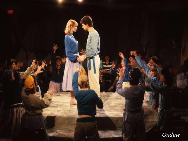 Two actors stand holding hands on a circular stage, surrounded by a group of performers with raised arms. The scene is lit by a spotlight, creating a dramatic atmosphere. The audience is seated around the stage.
