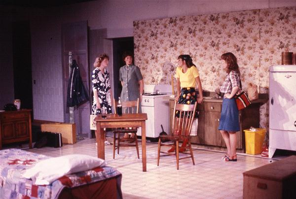 Four people are on a stage set resembling a kitchen with floral wallpaper. The group includes three women in various outfits standing near a table and a man near a fridge. A bed is partially visible in the foreground.