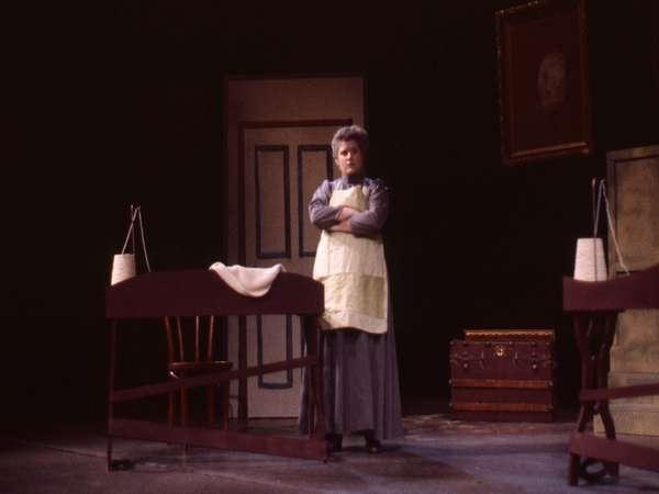 A person in period costume stands on stage with arms crossed. Behind them is a door and a framed portrait. Two cradles are visible, along with a trunk and a wooden chair. The scene has a dramatic, theatrical atmosphere.