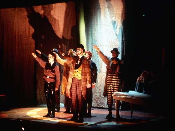 A group of performers on stage, dressed in vintage costumes, strike theatrical poses with arms outstretched. They are illuminated by dramatic lighting against a backdrop of textured curtains.
