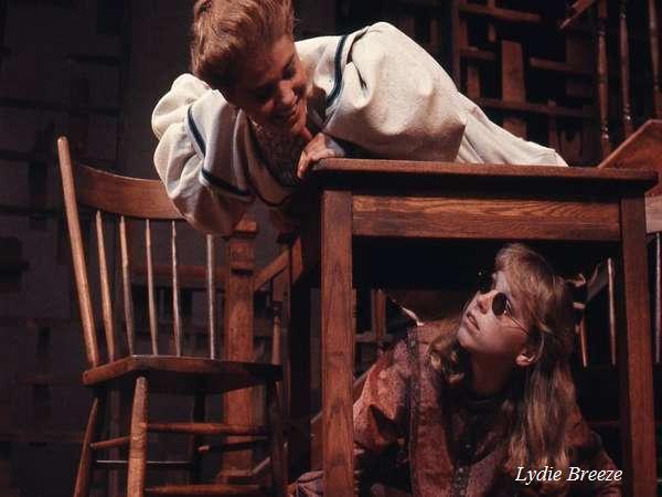 Two actors on stage; one leans over a table, wearing a white garment and smiling, while the other sits underneath, wearing glasses and gazing upward. Props and wooden furniture surround them, creating a theatrical setting. "Lydie Breeze" is visible.