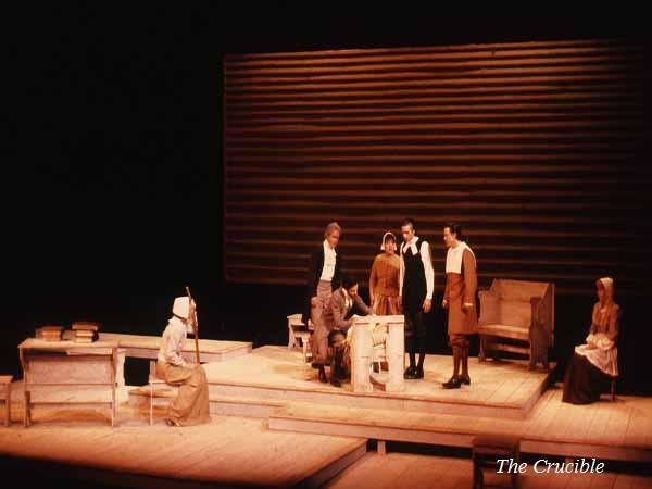 Stage performance of "The Crucible" with actors in period costumes on a wooden set. One actor is sitting in a chair, others stand nearby, and one is seated to the side. A minimalist backdrop sets the scene.