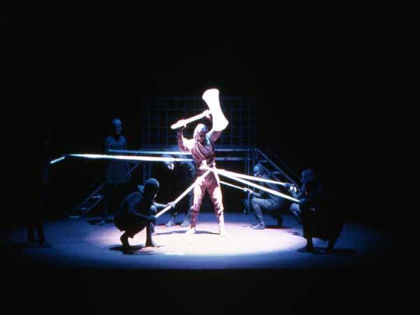 A theatrical performance with actors in dark outfits and masks performing under dim lighting. A central figure raises an object above their head, while others hold glowing lines, creating a dynamic and mysterious scene onstage.