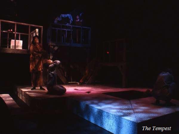 A dimly lit stage scene from a theater production of "The Tempest." Several actors are positioned on a raised platform and below it, partially obscured in shadow. The atmosphere is mysterious and dramatic.