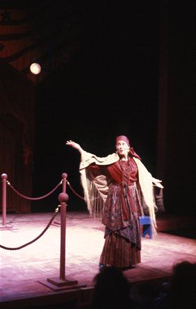 A person wearing a patterned dress, shawl, and headscarf performs on stage. They gesture with their right arm raised. The stage is dimly lit with a spotlight, and there are ropes and posts in the background.