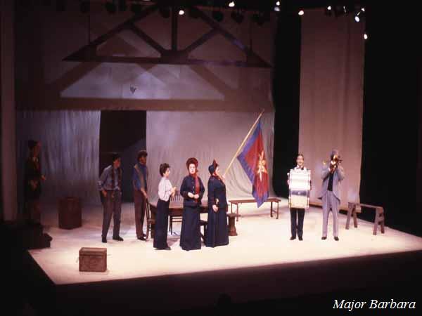 A stage performance of "Major Barbara" with actors in period costumes. The set includes minimalist props, a backdrop with beams, and a large flag. The scene is illuminated by stage lights.