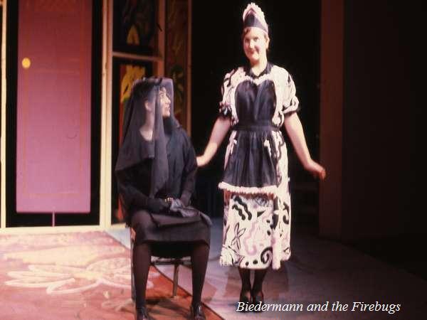 Scene from a stage play with two actors in costume. One is seated wearing a dark outfit with a veil, while the other stands smiling in a black and white dress with a patterned apron. The text "Biedermann and the Firebugs" appears in the corner.