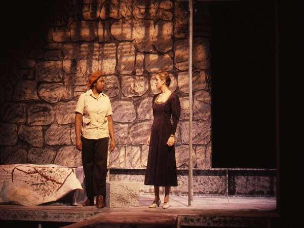 Two actors on stage perform against a stone wall backdrop. One wears a light shirt, dark pants, and a hat, while the other wears a long dark dress. A draped object is on the left. The lighting is warm and dim, creating a dramatic atmosphere.