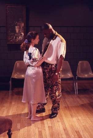 A man and woman stand closely on a wooden floor. The woman wears a light dress and the man wears a white shirt with patterned pants. They are gazing at each other in a dimly lit room with plastic chairs in the background.