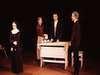 Four people are on stage in a dramatic scene. One stands off to the side in a dark dress, while the other three are gathered around a wooden table. The background is dimly lit, focusing attention on the characters.
