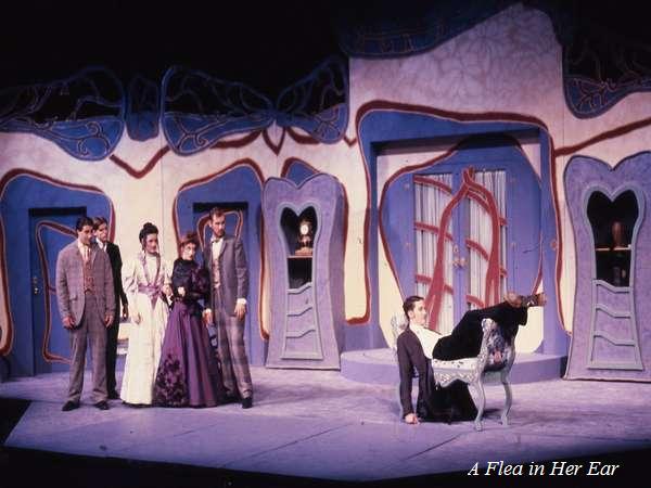 A theatrical performance of "A Flea in Her Ear," featuring five actors on stage in period costumes. The set is whimsically designed with blue and white doors and a curved arch. One actor is dramatically seated in the foreground.