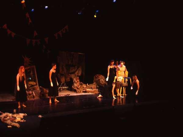 A group of performers in black costumes are on a dimly lit stage with cluttered props, including flags and an antique mirror. They appear engaged in a theatrical performance with dramatic lighting.