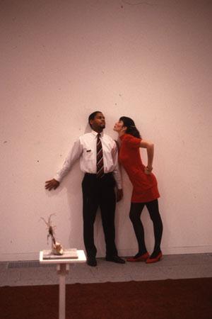 A man in a white shirt and tie stands against a wall. A woman in a red dress leans towards him with puckered lips. There is a small sculpture on a pedestal in the foreground.