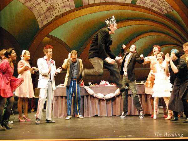 A lively stage performance with a group of people in colorful costumes. One person is jumping high with a crown on their head, while others around them clapping and cheering. The backdrop is an ornate, abstract design.