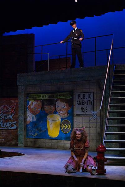 A dimly lit stage with a young woman in a tattered dress sitting near a fire hydrant, clutching a stuffed rabbit. Above, a man in a police uniform stands guard on a higher level. The backdrop features a colorful advertisement and a sign reading "Public Amenity No. 9.
