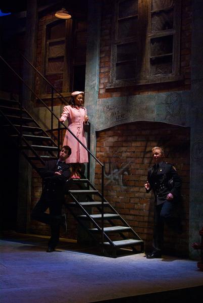 Three people are on a dimly lit stage resembling a brick alleyway. A woman stands on a stairway, wearing a pink coat and hat. Two men, dressed in dark clothing, stand leaning against the lower part of the staircase, looking upwards.