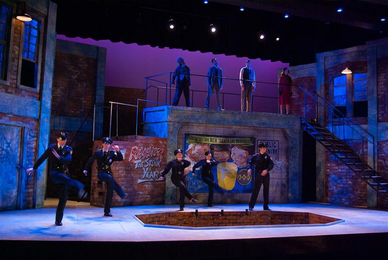 A stage scene features actors dressed as police officers performing a synchronized dance. The backdrop shows a graffiti-style mural with text and images. Additional actors stand on an elevated platform in the background. The lighting is vivid and colorful.