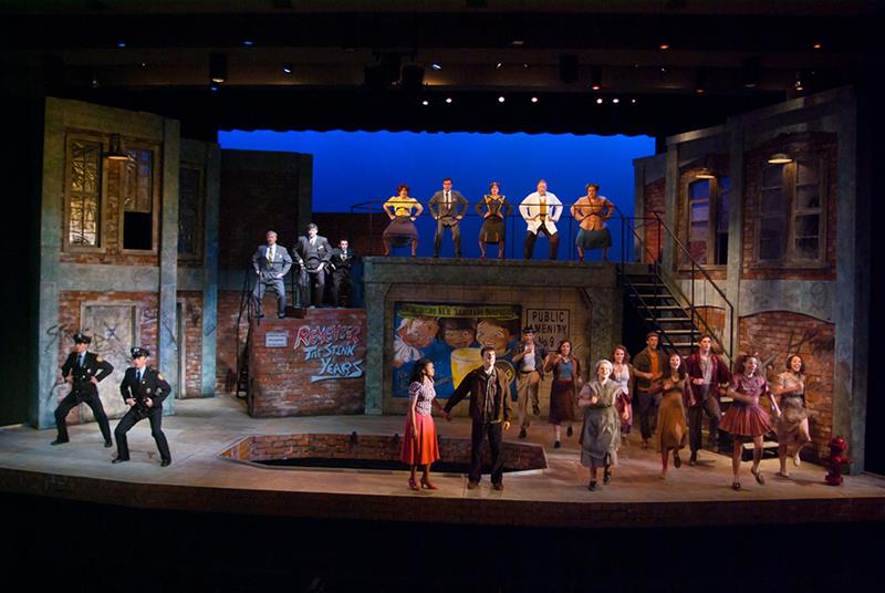 A stage production with a group of performers dressed in early to mid-20th century attire. The set features urban brick buildings and stairs. People are singing or acting in various poses, suggesting energy and emotion in the scene.