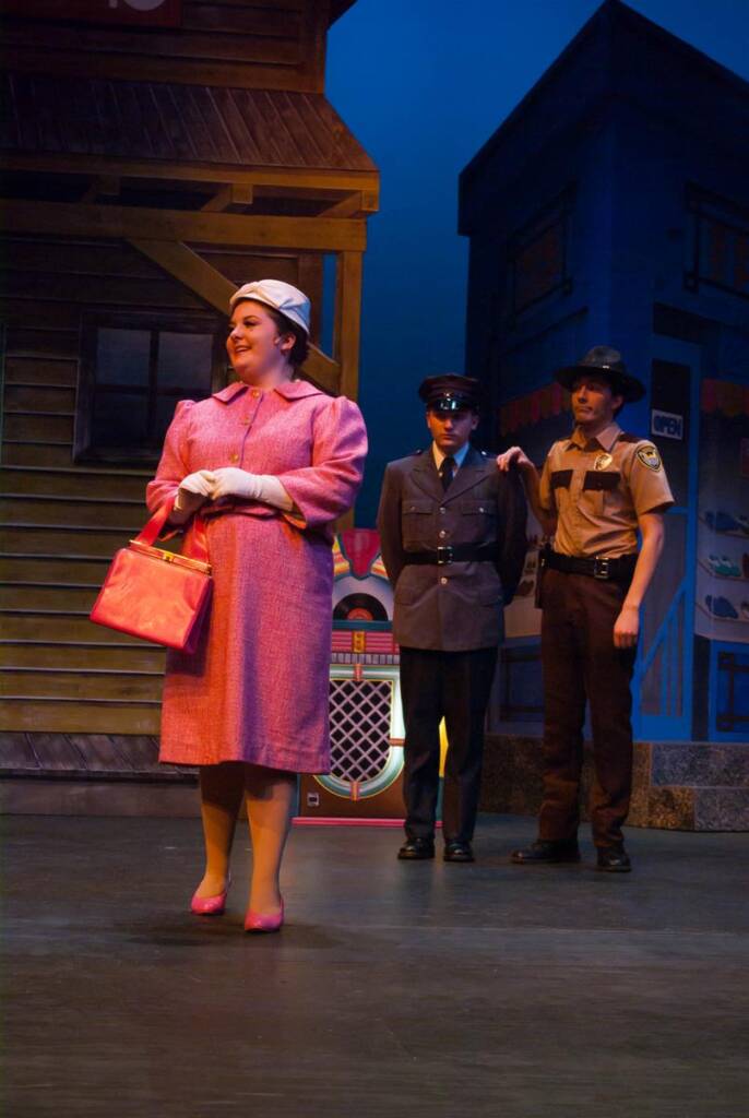 A person in a pink outfit stands smiling on stage, holding a matching purse. Two uniformed guards are behind, one in sunglasses. The backdrop resembles a town setting with buildings. The lighting is bright, giving a theatrical ambiance.