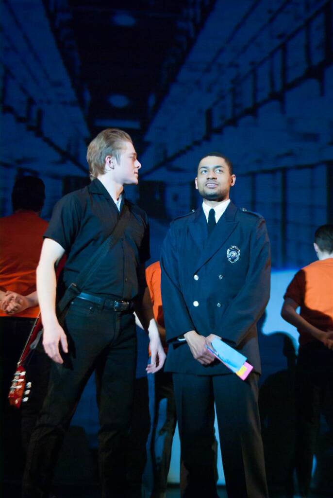 Two men perform on stage; one wears a dark shirt with a guitar strap, the other a security uniform. They are in front of a backdrop resembling a corridor. Other performers in orange outfits are visible in the background.