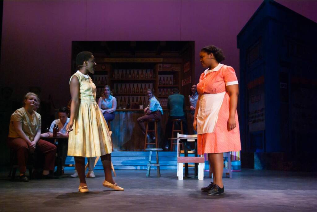 Two performers are on stage in a scene. One wears a yellow dress and the other a pink dress with an apron. They face each other with a bar setting and seated individuals in the background. The lighting is focused on them.
