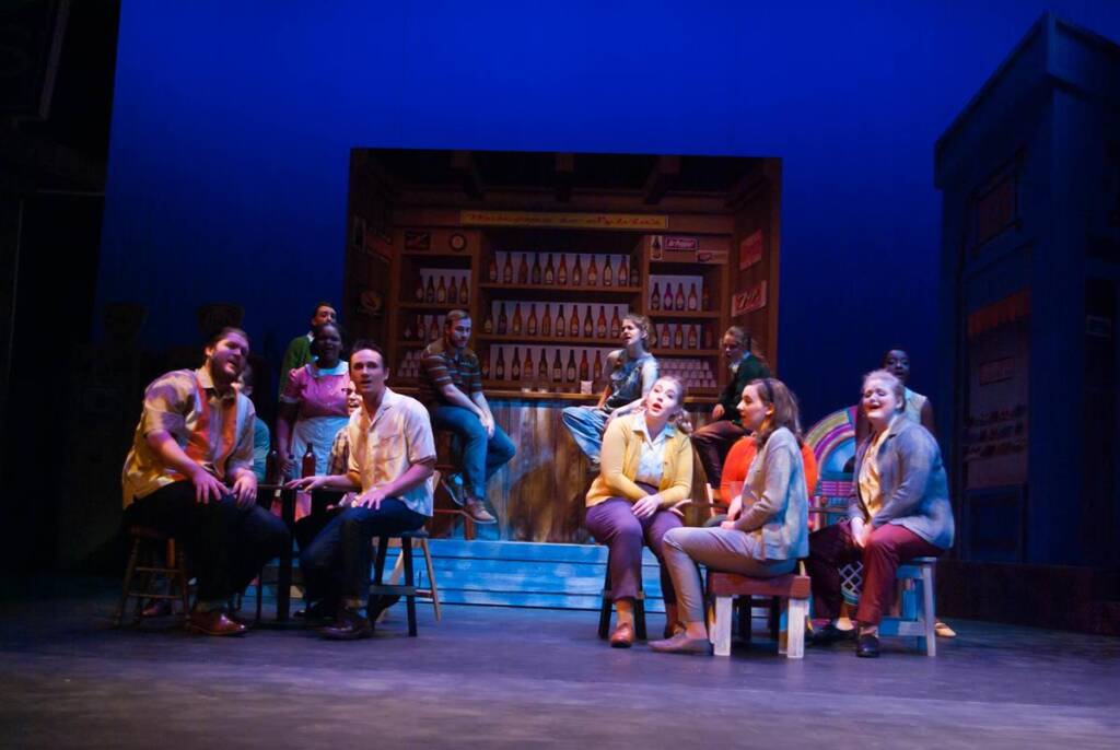 A group of performers on stage, sitting and standing around a bar set. The stage is dimly lit with a blue background, and bottles are displayed on shelves in the set. The actors are wearing casual clothing and appear to be engaged in conversation.