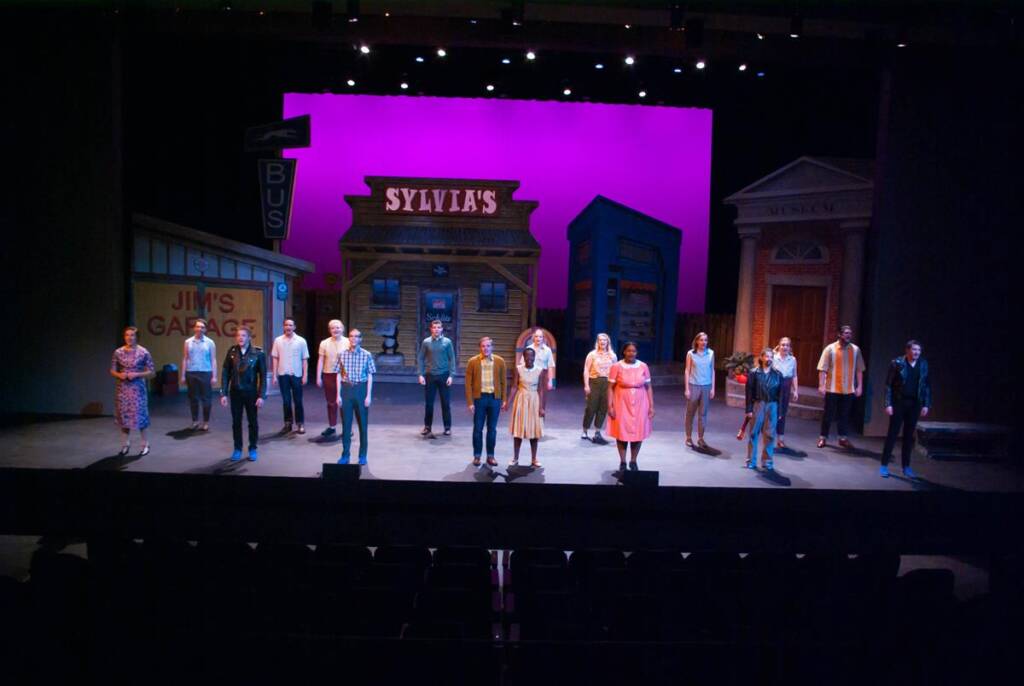 A group of actors perform on a stage with colorful building facades in the background, including signs reading "Sylvia's" and "Jim's Garage." The stage is lit with a purple backdrop.