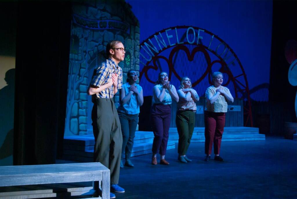A group of five actors performs on stage in front of a backdrop that reads "Tunnel of Love." They are standing in a line, with one in front gesturing animatedly. The stage is dimly lit with blue light.