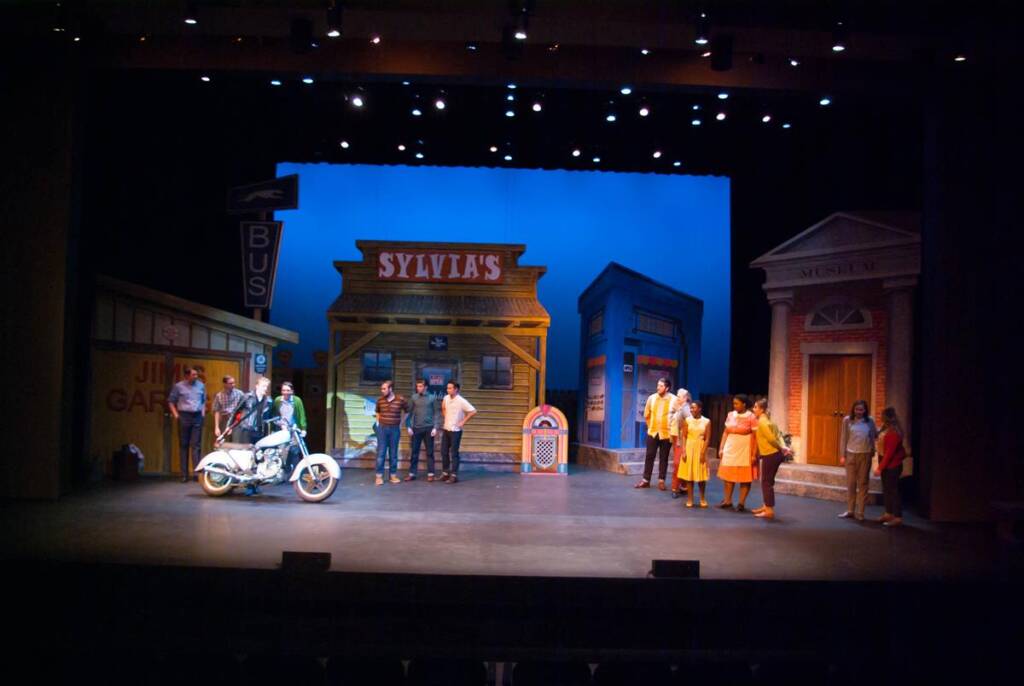A theater stage scene depicts a colorful street with storefronts labeled "Sylvia's" and "Jim's Garage." People are gathered in various groups, some near a motorcycle, under stage lights against a blue background.
