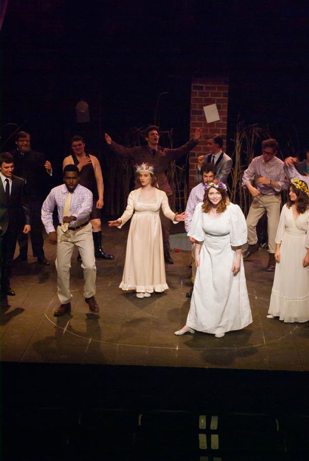 A group of people in costumes perform on stage. Two women in white dresses stand in the foreground with flower crowns. Others are dressed in various casual outfits, and a man in a suit is on the left. The background is dark with scattered props.