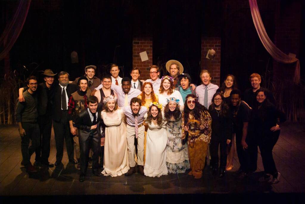 A group of twenty-four people in costume poses on a stage. The background features brick pillars and flowing drapes. Some individuals are dressed in wedding attire, while others wear casual or period clothing. Everyone is smiling and standing closely together.