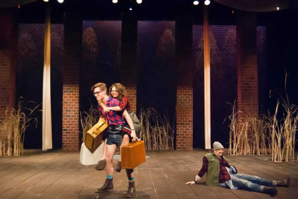 A stage performance features a person carrying another on their back, both holding suitcases. A third person is seated on the wooden floor in the background. The set includes tall grass decorations and brick wall panels.
